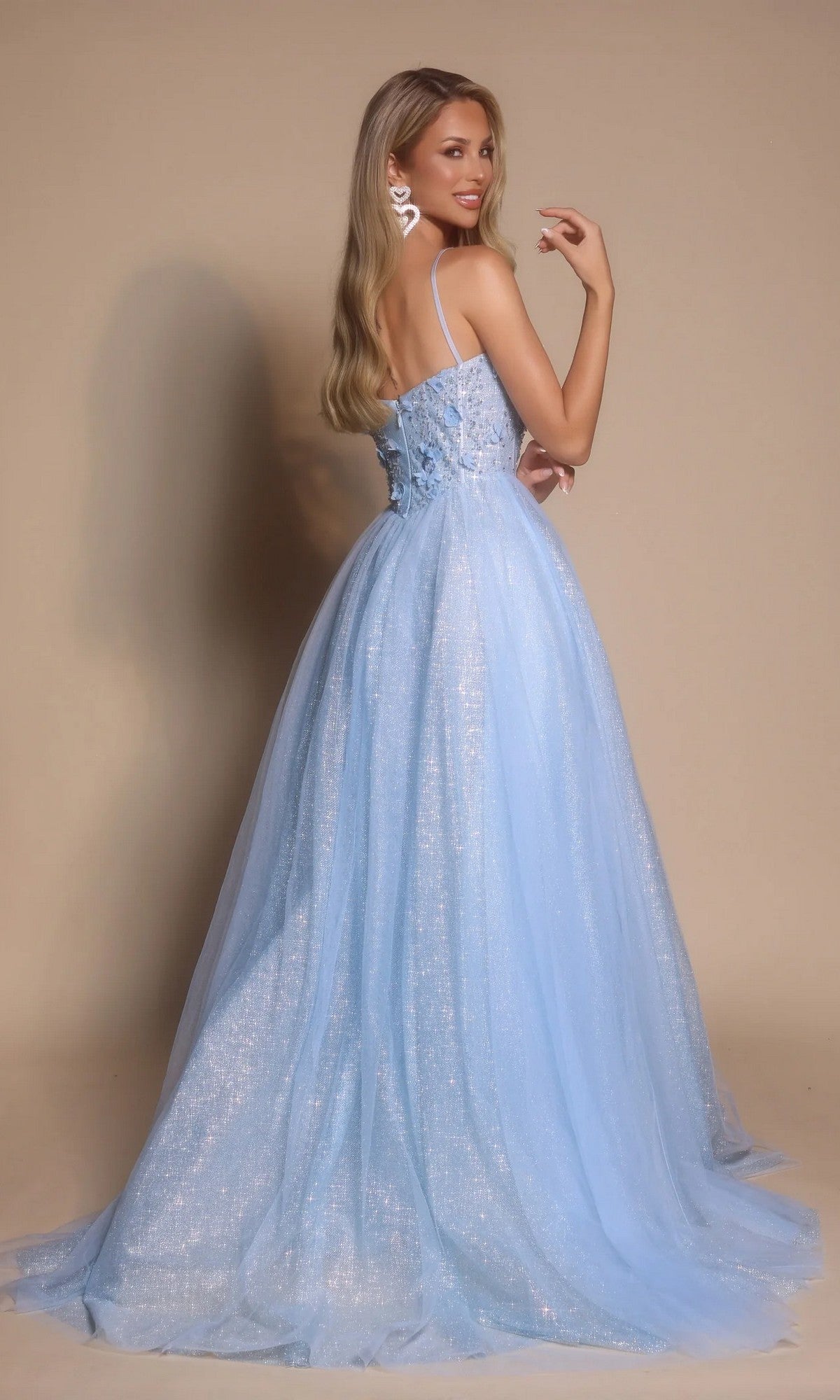 A woman in the Portia and Scarlett PS26146 Glitter Prom Ball Gown poses with her back turned, glancing over her shoulder. The light blue floor-length dress features glitter tulle and a boned-corset bodice against a beige background.